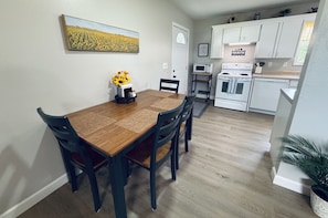 A dining area with a wooden table and chairs, a kitchen with white cabinets and appliances, and a painting on the wall.