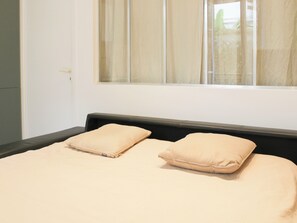 Ein Bett mit zwei beigen Kissen, einem schwarzen Kopfteil und einem Fenster mit transparenten Vorhängen.