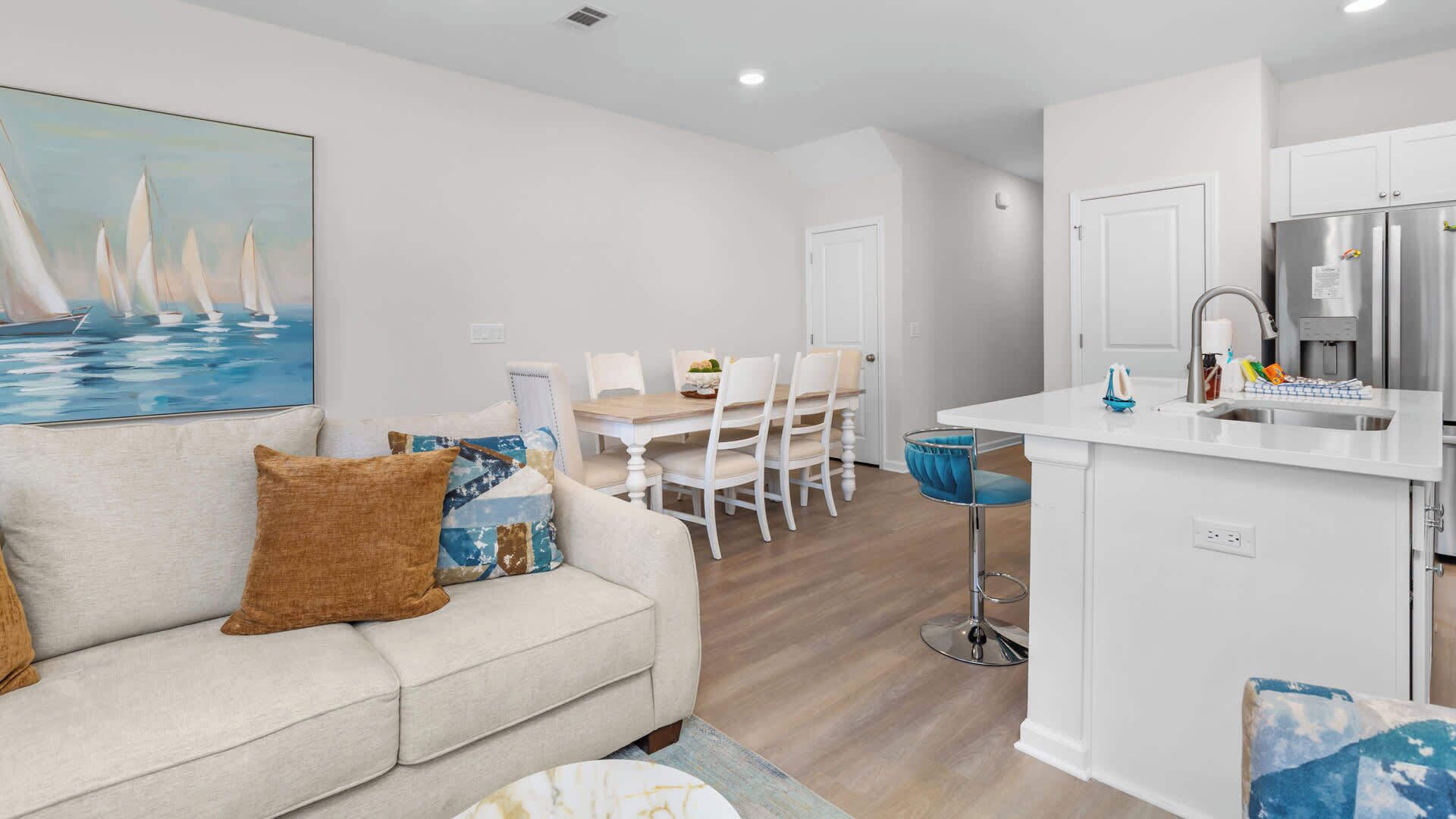 A modern living room with a sofa, a dining table with chairs, a kitchen with a sink and refrigerator, and a painting on the wall.