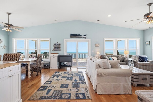 A spacious living room with a large window, a ceiling fan, and a view of the ocean. - Knotty in the Kitchen: Oceanfront Near Park (Oak Island)