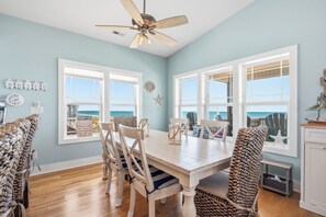 A dining area with a white table and chairs, a ceiling fan, and large windows with a view of the ocean. - Knotty in the Kitchen: Oceanfront Near Park (Oak Island)