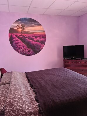 Ein Schlafzimmer mit einem Bett, einem Fernseher, einem Schminktisch und einem runden Wandbild, das einen Sonnenuntergang über Lavendelfeldern zeigt.