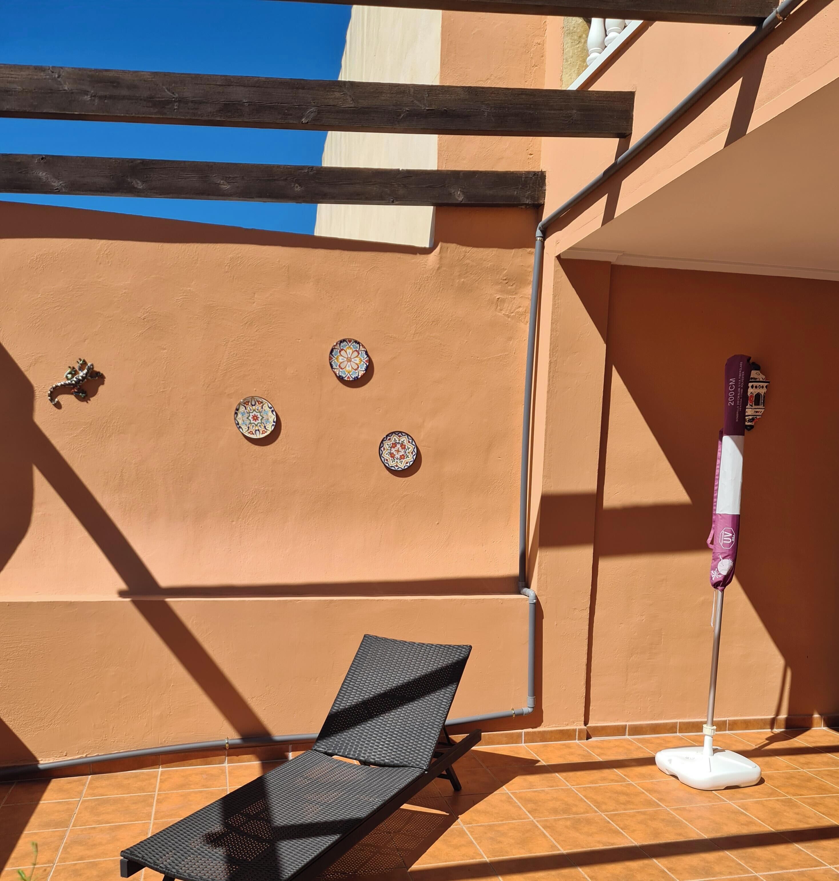 Un espacio al aire libre iluminado por el sol con una silla de salón negra, un trapeador y platos decorativos en una pared.
