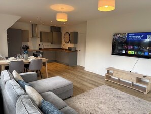 A modern living room with a grey sofa, wooden flooring, a dining area with chairs, a kitchen in the background, and a TV displaying a booking offer.