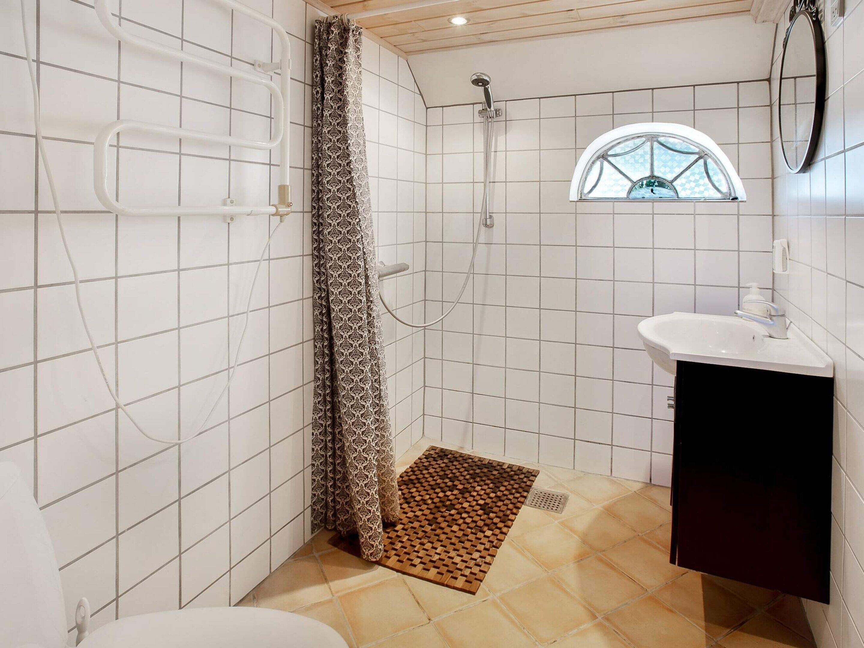 A bathroom with white tiled walls, a shower with a patterned curtain, a small window, and a sink with a black cabinet.