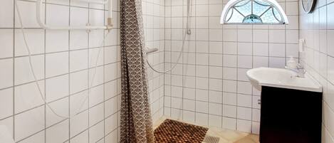 A bathroom with white tiled walls, a shower with a patterned curtain, a small window, and a sink with a black cabinet.