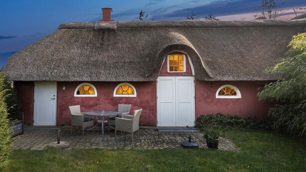 Uma casa com telhado de palha e porta branca, um pequeno pátio com mesa e cadeiras, e um jardim.