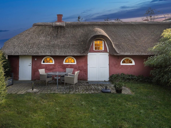 Ein Haus mit einem Strohdach, einer weißen Tür, einer kleinen Terrasse mit Tisch und Stühlen sowie einem Garten.