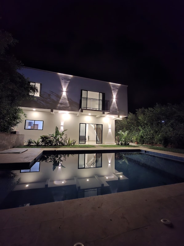 A modern house with a swimming pool, a balcony, and a well-lit exterior.