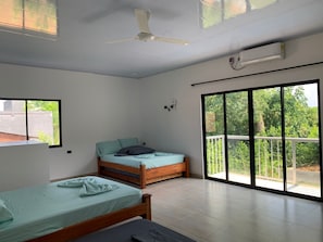 A bedroom with two beds, a ceiling fan, an air conditioning unit, and a large sliding glass door leading to a balcony with a view of greenery.