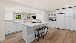 A modern kitchen with white cabinetry, a central island, wooden stools, and recessed ceiling lights. - Hidden Harbor Getaway-Sunrise Views-Private Beach Access (Gig Harbor)