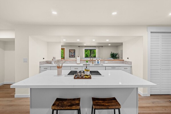 A modern kitchen with a large island, white cabinetry, and a view of greenery outside.