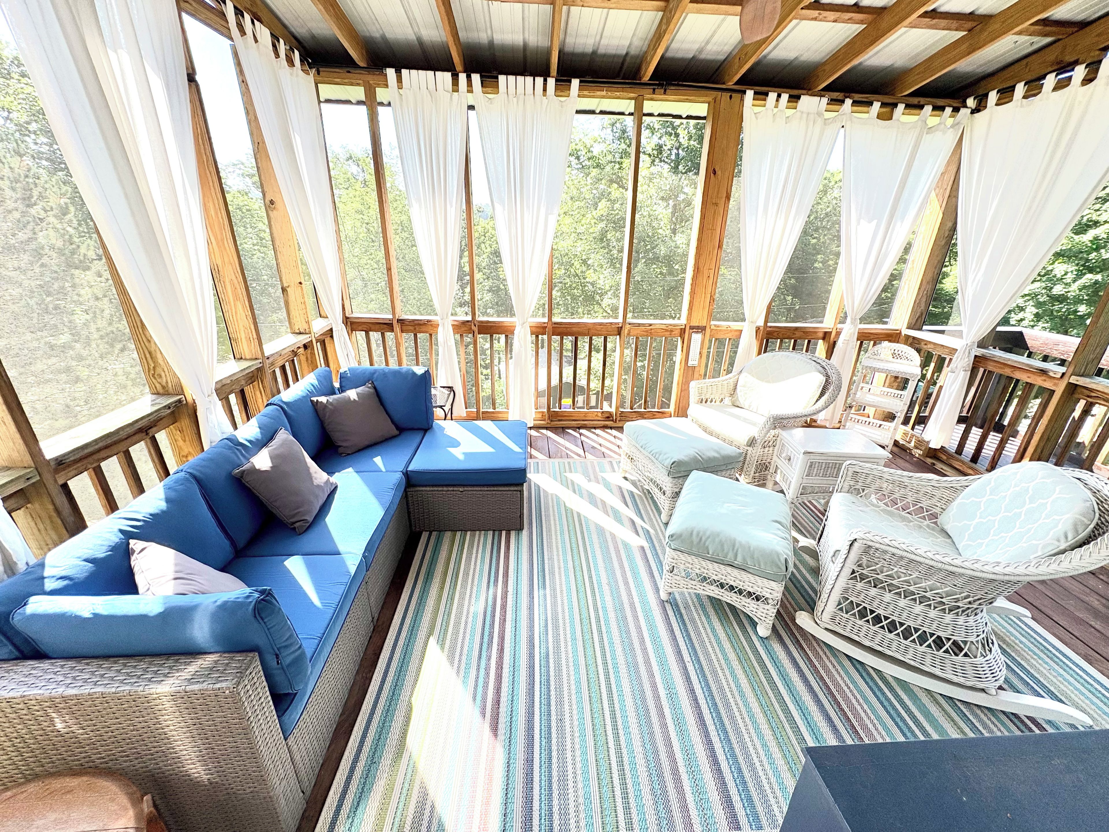 Un espace extérieur aménagé avec des sièges, abrité par un toit, comprenant un canapé bleu, des fauteuils en osier et un tapis à rayures.