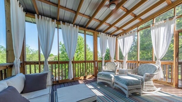 A covered outdoor seating area with wicker furniture, a cushioned bench, and a view of greenery.