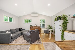 A modern living room with a sectional sofa, a small dining table, a piano, and a kitchen area with a refrigerator and plants.