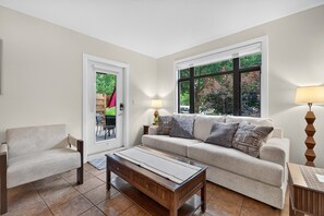 Ein Wohnzimmer mit einer Couch, einem Sessel, einem Couchtisch und einer Lampe. Ein Fenster bietet Blick auf Bäume, und eine Tür führt nach draußen.