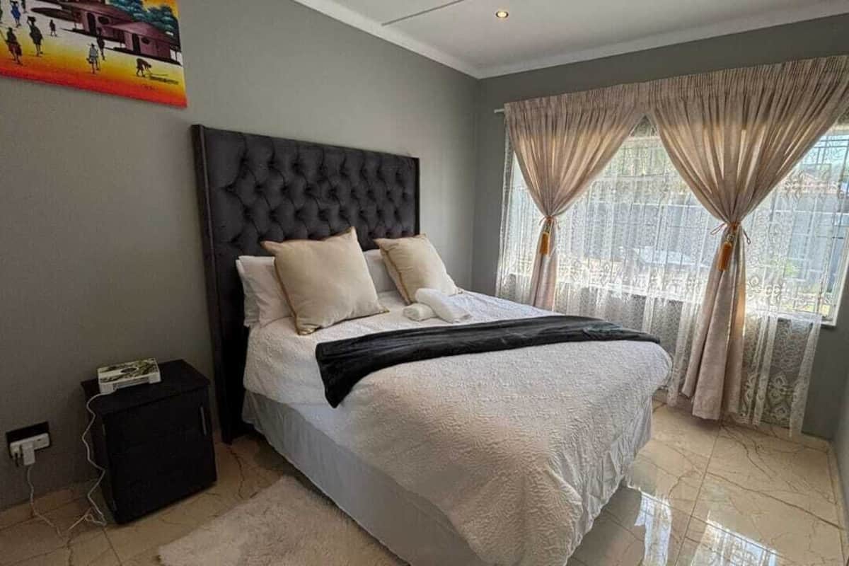 Ein Schlafzimmer mit einer gepolsterten Kopfstütze, einem Bett mit weißen Leinentextilien, einer schwarzen Tagesdecke und einem Fenster mit durchsichtigen Vorhängen.