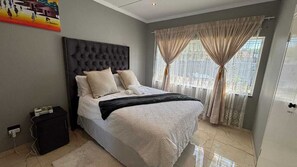 Un dormitorio con un cabecero capitonado, una cama con sábanas blancas, un plaid negro y una ventana con cortinas de gasa.