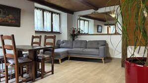 A room with a wooden dining table, four chairs, a grey sofa, a red potted plant, and a wooden ceiling.