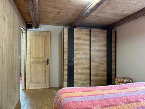 A bedroom with a wooden ceiling, a wooden wardrobe, a bed with a striped blanket, and a small floral bag on the floor.