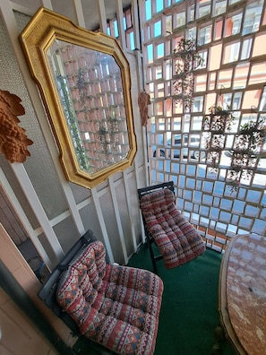 Ein Zimmer mit einem großen, verzierten Spiegel, zwei gemusterten Hocker, einem runden Tisch und Blick durch ein Gitterfenster.