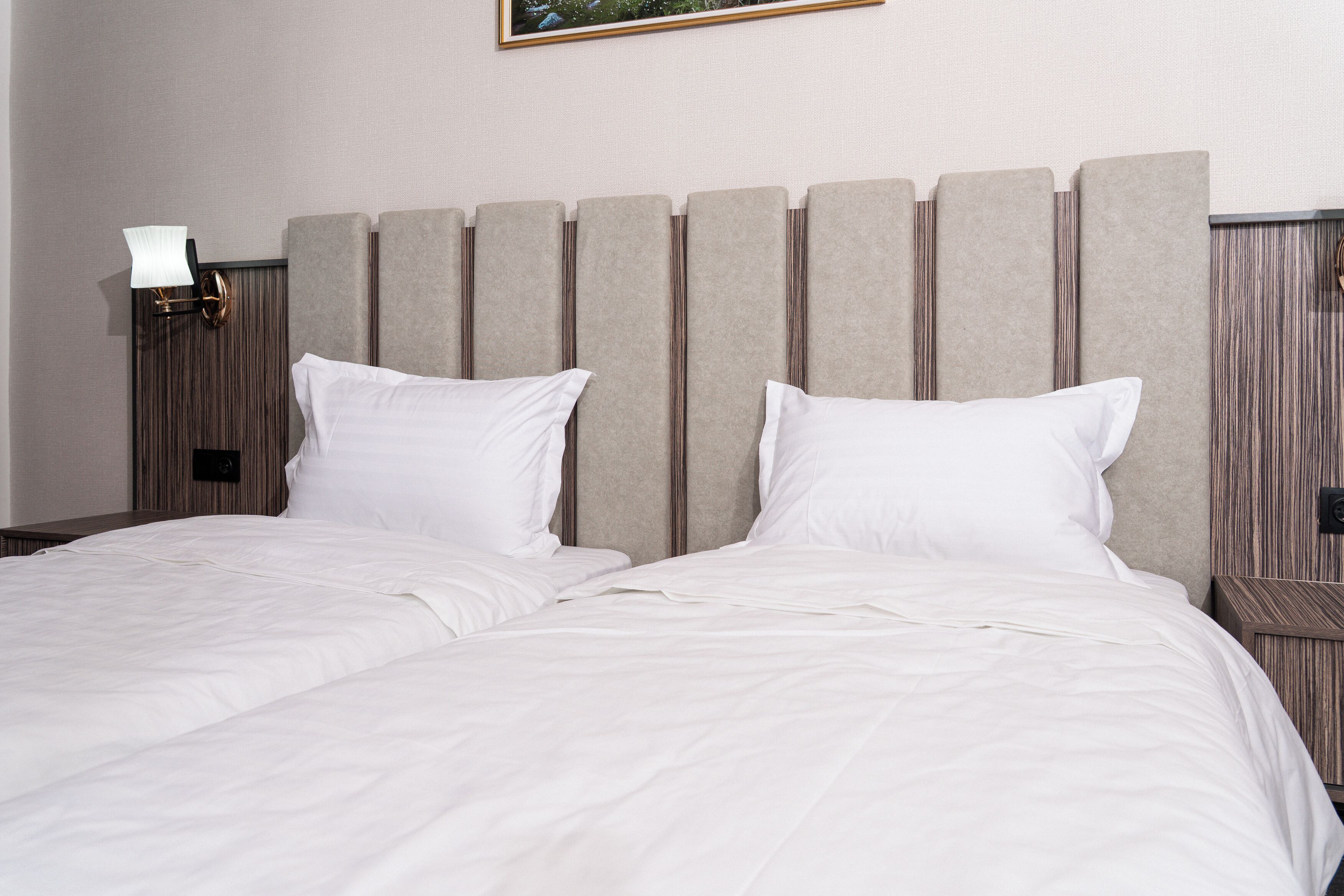 Een netjes opgemaakt bed met witte lakens, een hoofdbord met verticale panelen, en een wandmontage nachtkastje met een lamp.