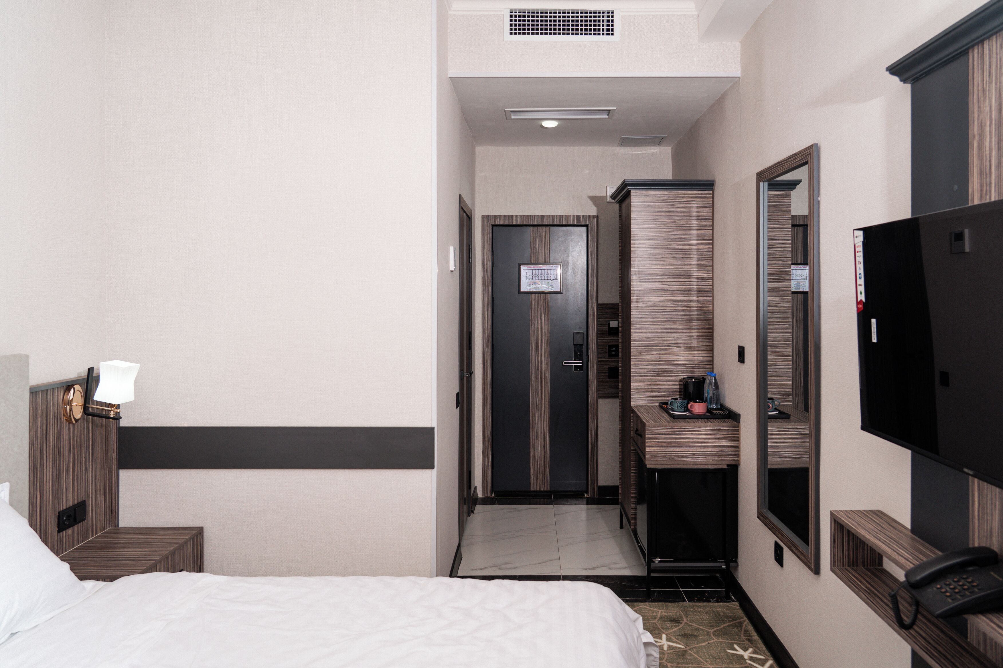 A hotel room with a bed, a bedside table with a lamp, a mirror, a TV, and a bathroom visible through an open door.