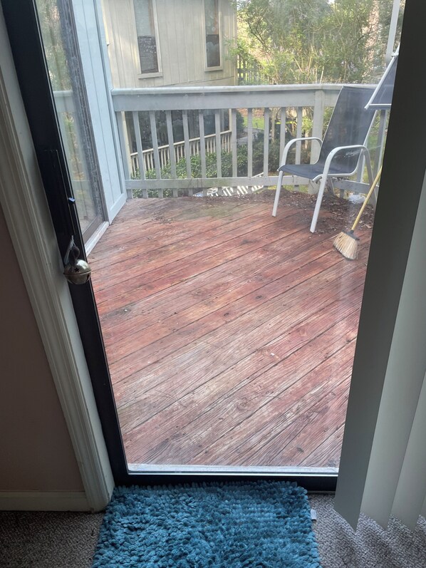 Una terraza de madera con barandilla blanca, un felpudo azul en la entrada y una escoba apoyada contra la barandilla.