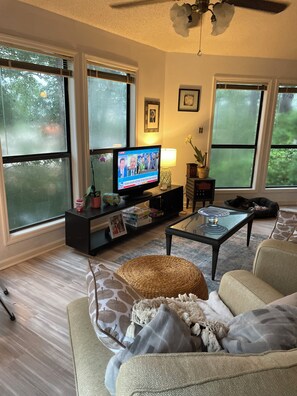 A living room with a television displaying a news broadcast, a sofa, a coffee table, and large windows. - Cozy 2-bedroom house in amazing Myrtle Beach with AC (Myrtle Beach)