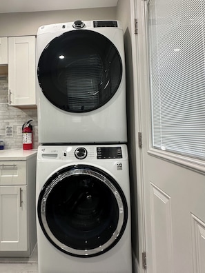 A stacked washer and dryer unit in a kitchen with white cabinets and a window with blinds. - Game Day Haven — 8 min Walk to AT&T Stadium —Sleeps 10 (Arlington)
