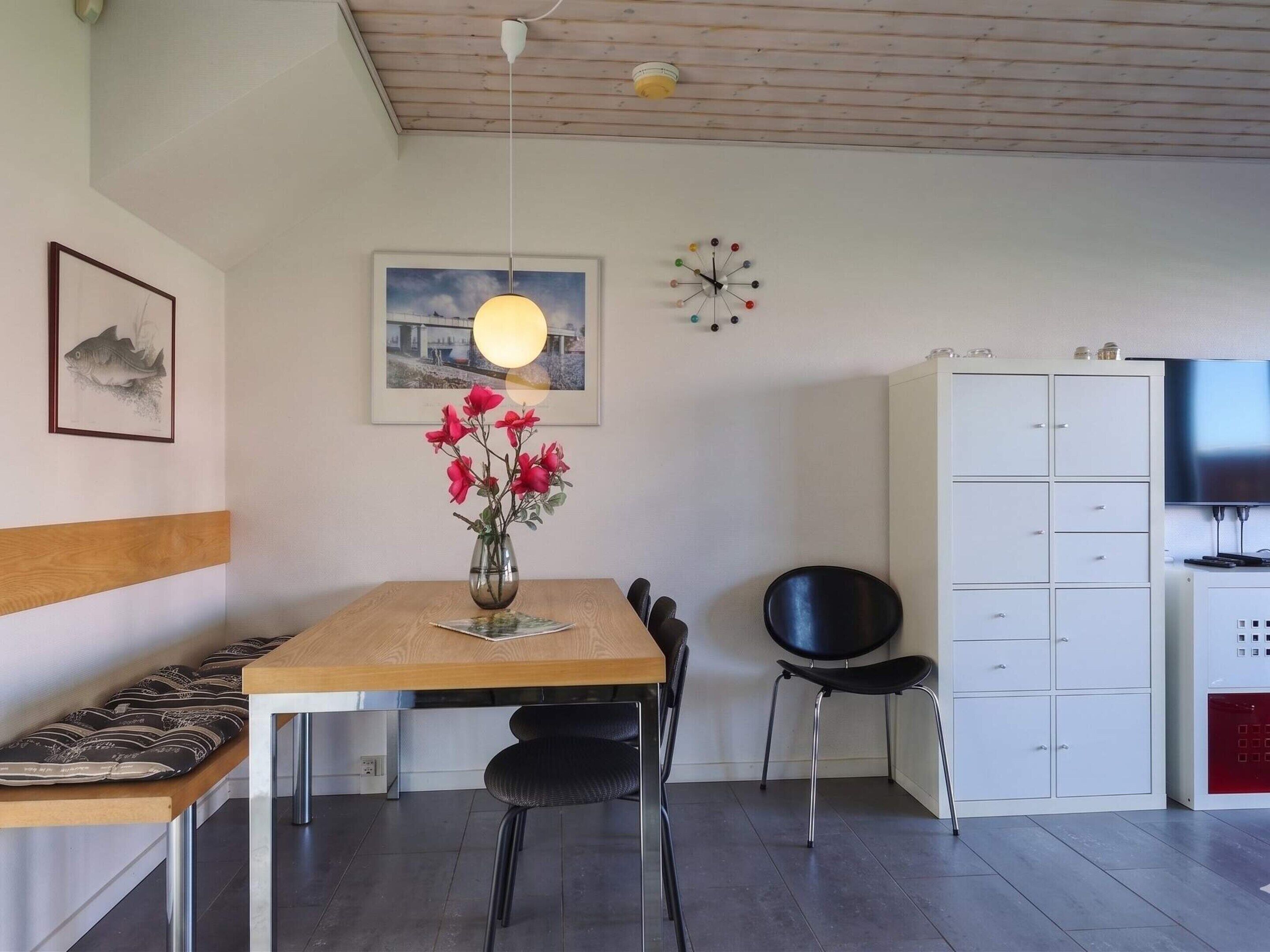 A modern room with a wooden dining table, black chairs, a vase with red flowers, a wall clock, and a television.