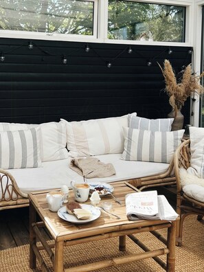 Ein gemütlicher Sitzbereich mit einer Couch, einem Holztisch mit Frühstücksleckereien und einem Korbstuhl.