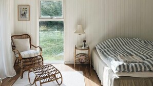 A room with a bed, a wicker chair, a small table with a lamp, a window with a view of greenery, and a framed picture on the wall. - Personal style in a home between Vig and Høve (Vig)