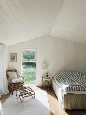 Ein Zimmer mit einem Bett, einem Korbstuhl, einem kleinen Tisch mit Lampe, einem Fenster mit Blick auf Grünflächen und einem Bilderrahmen an der Wand.