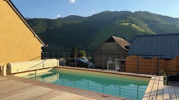 Une terrasse en bois avec une piscine, un bâtiment en pierre et une vue sur les montagnes en arrière-plan.