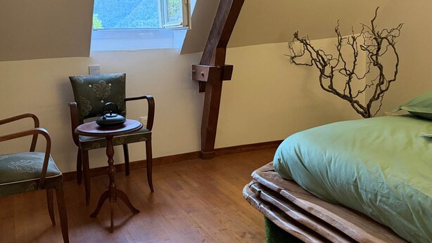 A bedroom with a sloped ceiling, a window with a view of mountains, a bed with a green cover, a chair with a decorative cushion, and a wooden floor. - Rental: Gazost Fountains (Gazost)