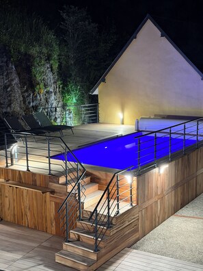 Piscina com deck de madeira e escadas, cercada por um edifício com fachada branca.
