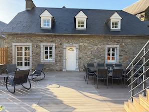 A stone building with a dark roof, a white door, and a staircase leading to a deck with outdoor seating.