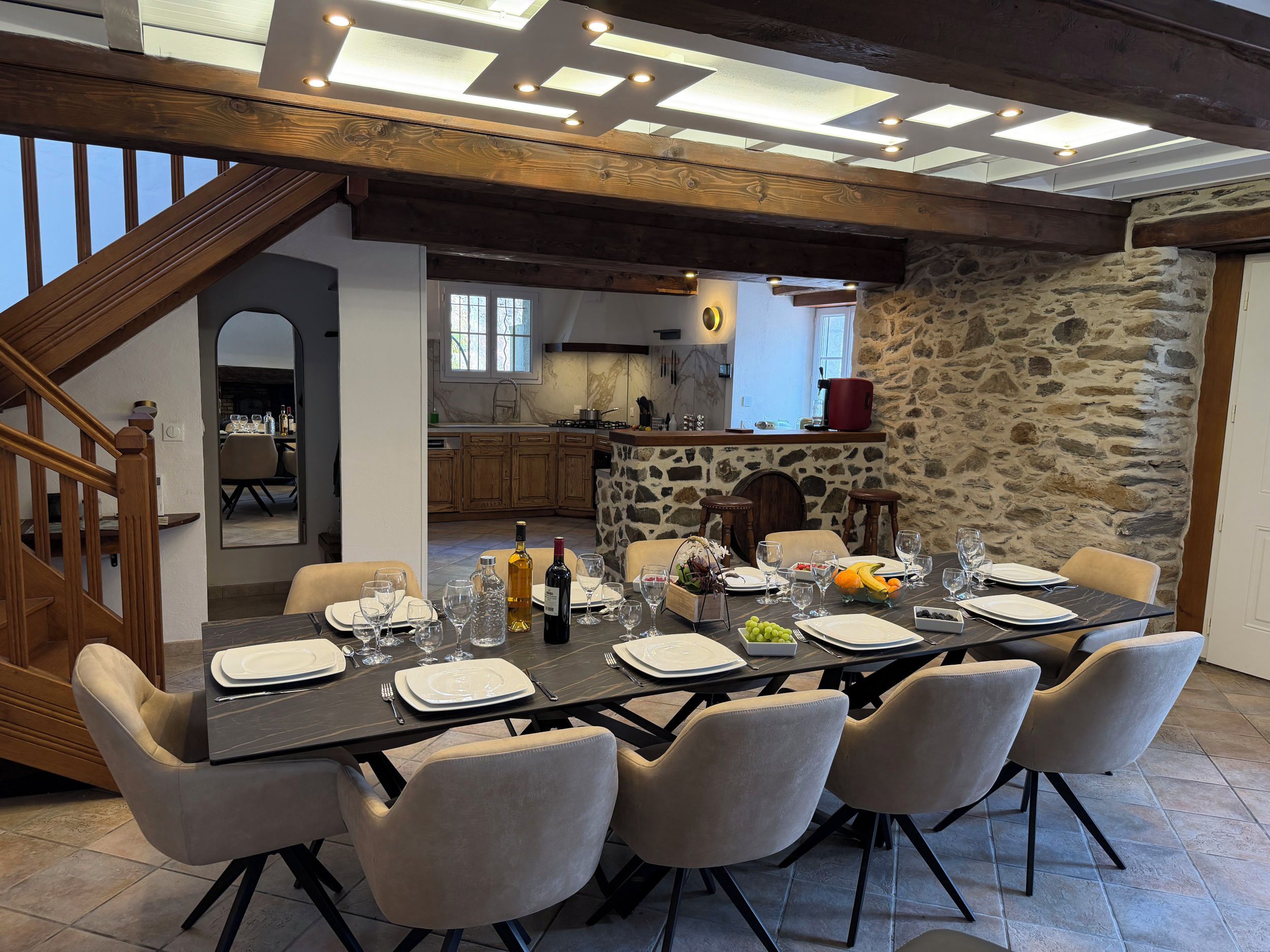 Un coin repas avec une grande table dressée pour six personnes, entourée de chaises, et une cuisine en arrière-plan.