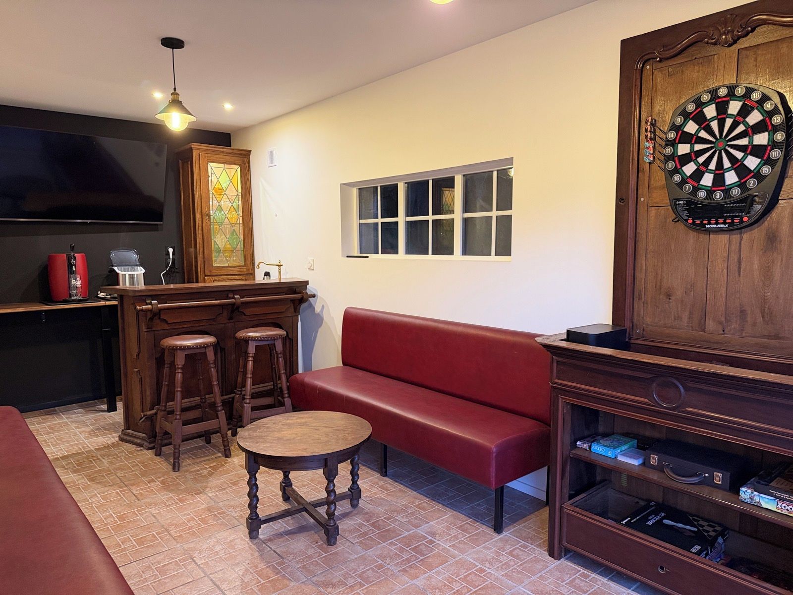 Une pièce avec un canapé rouge, un coin bar en bois, un jeu de fléchettes et un téléviseur fixé au mur.