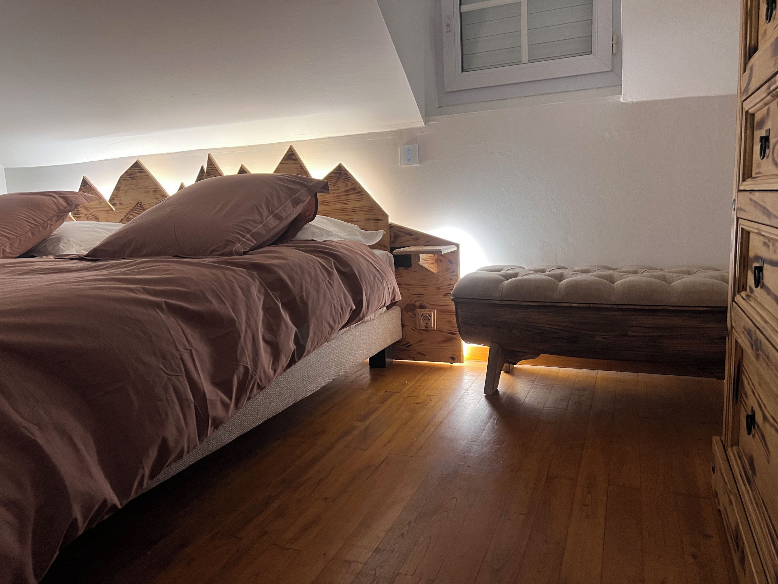 Een slaapkamer met een bed, nachtkastje, houten dressoir en een raam.