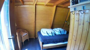 A loft-style bedroom with a wooden ceiling, a bed with a mattress and pillows, a small chair, and a shelf with items.