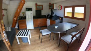 A compact kitchen with a refrigerator, microwave, and wooden cabinets. There is a dining table with chairs and a small bench. A clock is on the wall, and a window provides natural light.