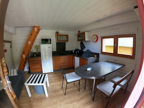 Une cuisine compacte équipée d’un réfrigérateur, d’un four à micro-ondes et de meubles de rangement en bois. On y trouve une table à manger avec des chaises et un petit banc. Une horloge est accrochée au mur, et une fenêtre laisse entrer la lumière naturelle.