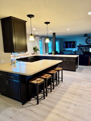 A modern kitchen with dark cabinetry, a central island with stools, pendant lighting, and a stone fireplace. - Hummingbird House: Relax and Connect in a Large Private Lakefront Home Sleeps 20 (Storm Lake)