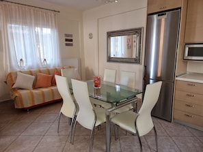 A modern kitchen with a stainless steel refrigerator, a glass dining table with white chairs, and a sofa with striped cushions.