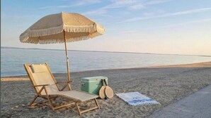 Ein Strandstuhl, ein Strohdachschirm, eine Kühlbox und ein Handtuch im Sand in der Nähe des Wassers.