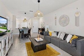 Un salon moderne avec un canapé gris, une table basse et un téléviseur posé sur un meuble. On aperçoit un coin repas avec une table et des chaises, ainsi qu’une cuisine au fond.