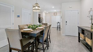 Uma área de refeições com uma mesa posta para quatro pessoas, rodeada por cadeiras, um lustre e vista para a cozinha.