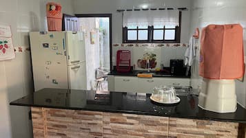 Cozinha com bancada preta, armários brancos, geladeira e janela com cortina de renda.